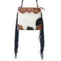 A western-style shoulder bag featuring a black and white cowhide body, black fringe hanging from the sides, and a tooled brown leather top panel with floral and scroll designs.
