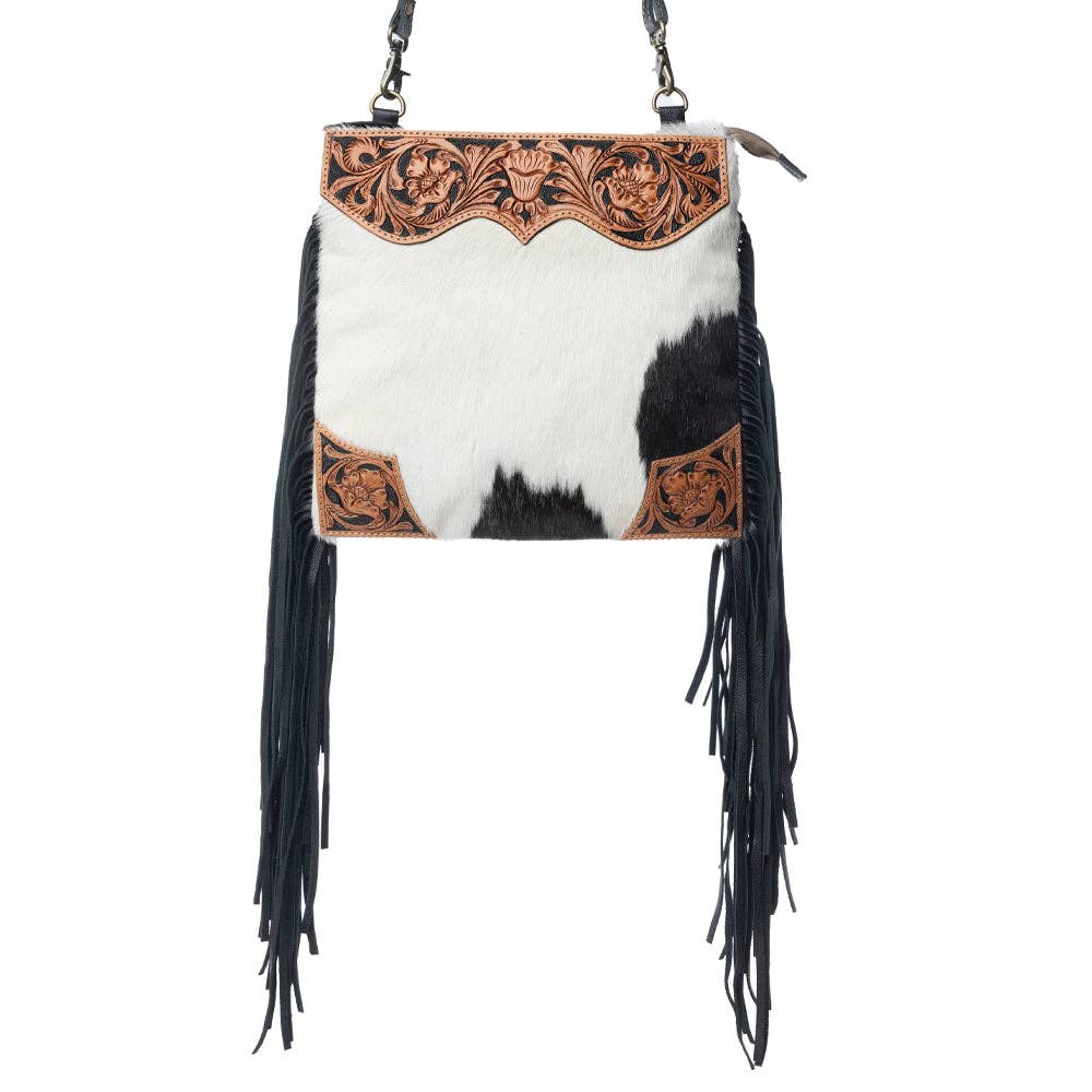 A western-style shoulder bag featuring a black and white cowhide body, black fringe hanging from the sides, and a tooled brown leather top panel with floral and scroll designs.